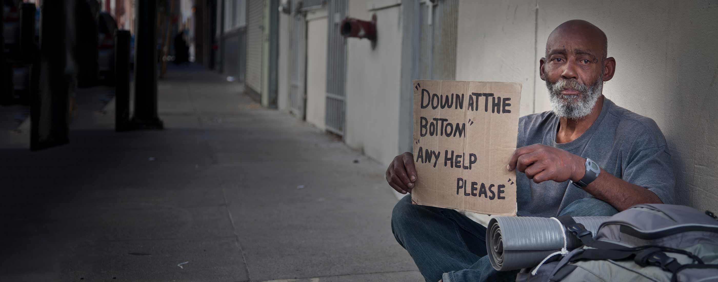 Photo of homeless man holding up sign that reads "Down at the bottom - Any help please"